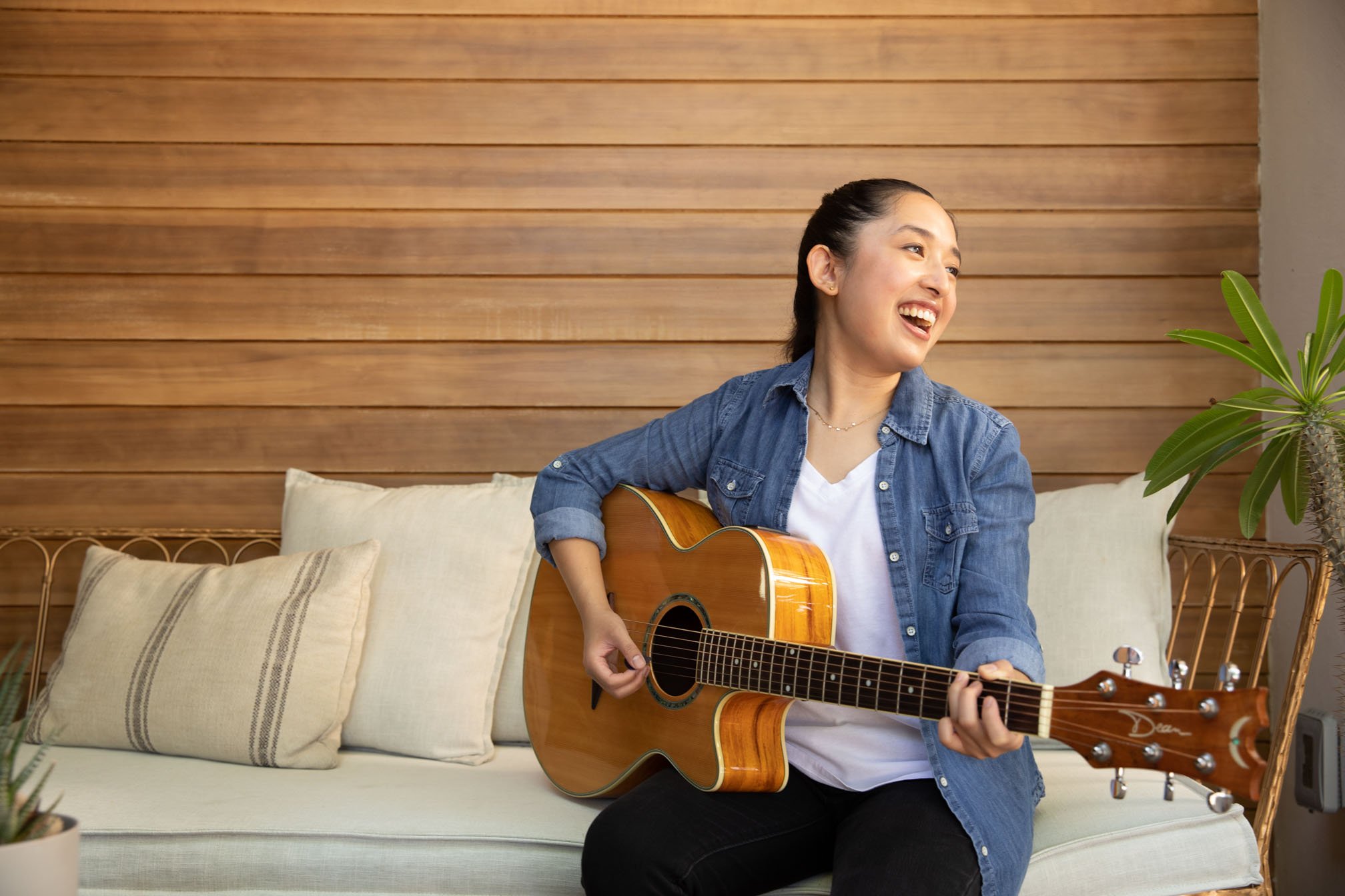 A woman with a guitar sits on a couch and smiles to someone off camera.