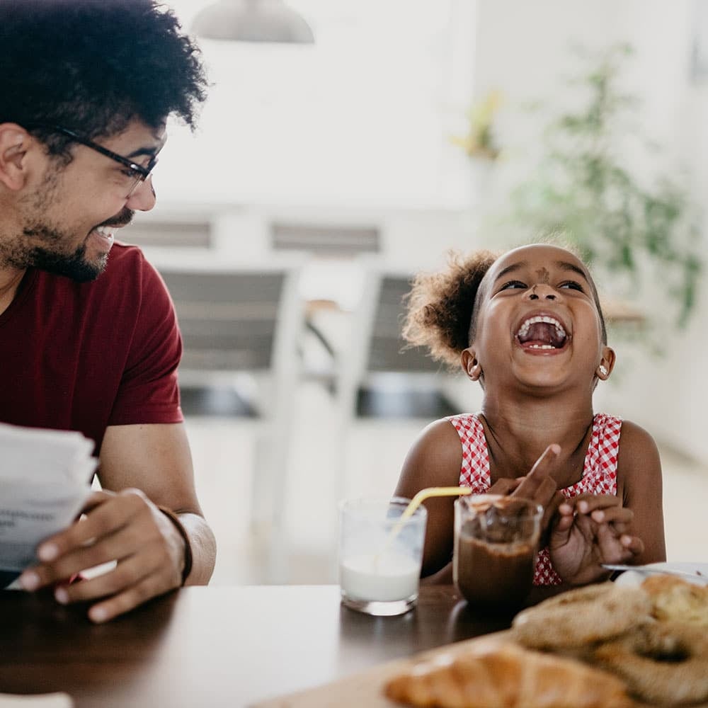 An image of a father and daughter enjoying pastries and milk at a table, laughing while the father reviews some papers.