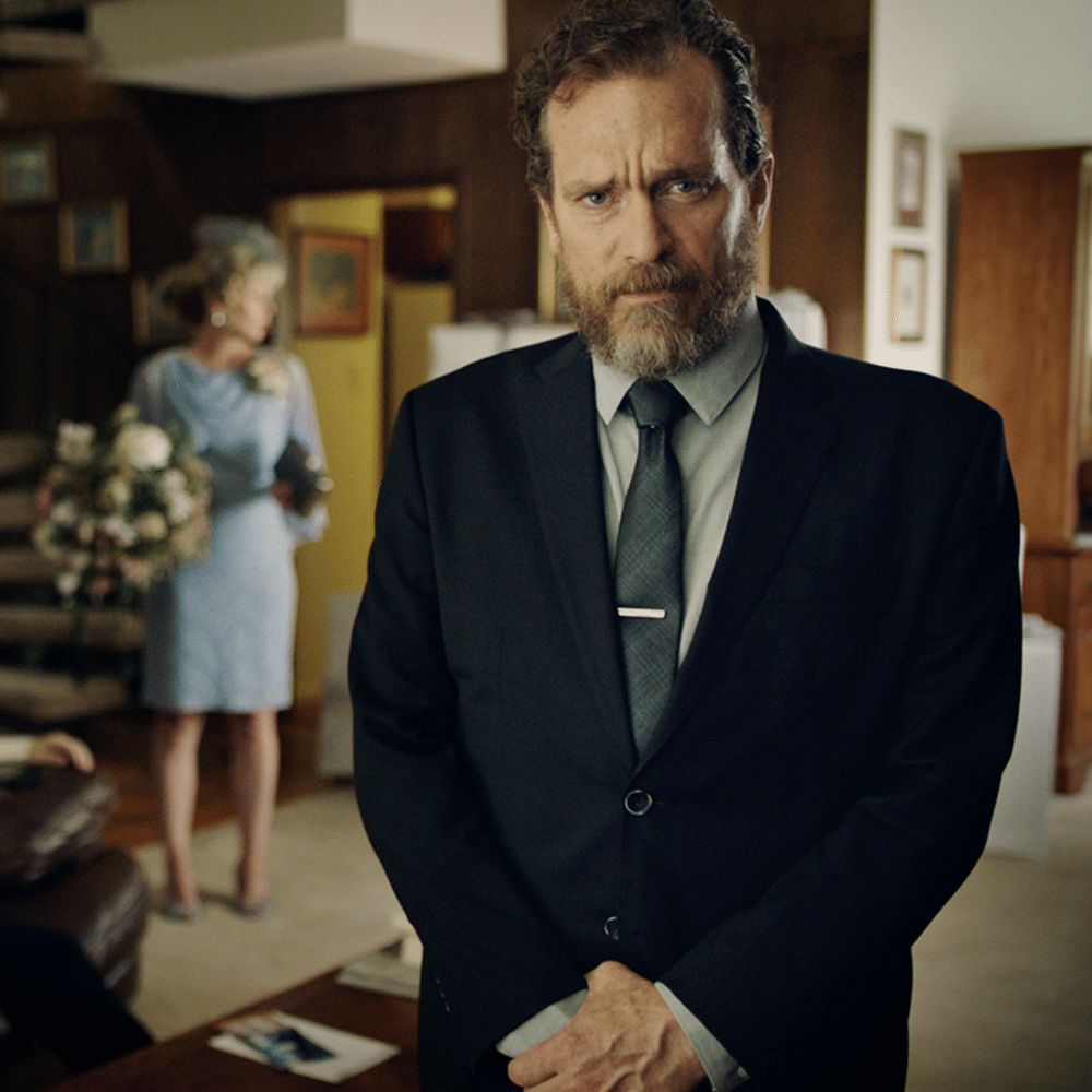 An image of a man in a suit, looking at the camera with a stern look, while someone looks impatient behind him.