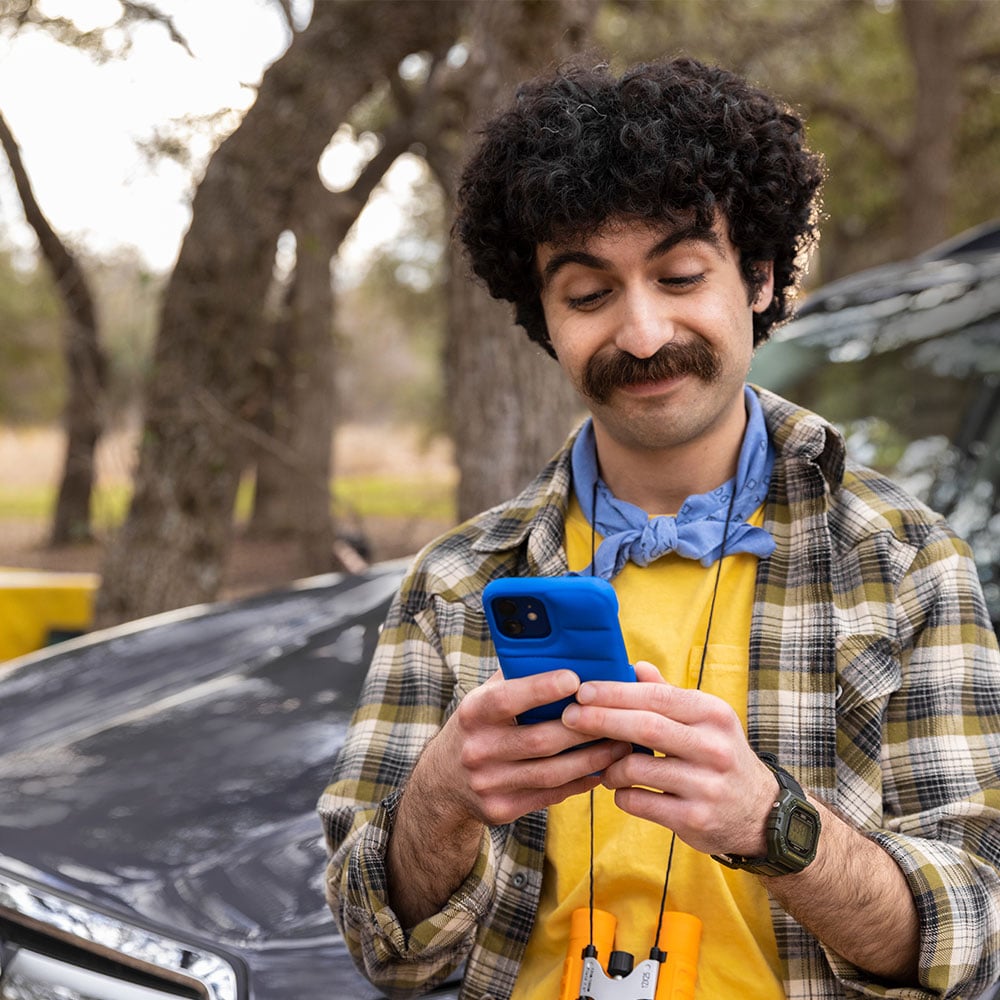 An image of a young man with a mustache, decked out with camping clear like a bandana and binoculars, looking at his phone as he stands next to his car.